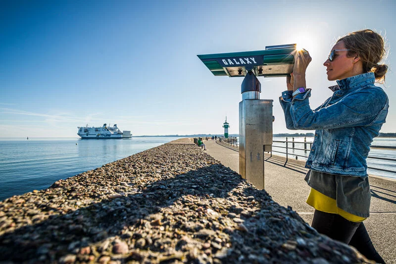 Eine Frau schaut aus einem Fernrohr auf der Nordermole in Travemünde. Im Hintergrund fährt ein Schiff.